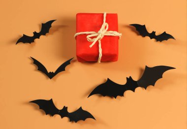 Kırmızı hediye kutusunun üstü ya da üstü turuncu arka planda siyah kağıt yarasalar ve fotokopi alanı ile kaplı. Cadılar Bayramı hediyesi kavramı.