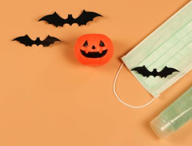 Cadılar Bayramı balkabağı, siyah kağıt yarasalar, tıbbi maske ve turuncu arka planda alkol dezenfektanı. Cadılar Bayramı, COVID-19 önleme ve yeni normal kavram.