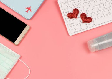 Bilgisayar klavyesinde, cep telefonunda, pasaport, tıbbi maske ve pembe arka planda dezenfektan jeli gibi kırmızı parıltılı kalplerin üst görüntüsü veya düz yatışı. Covid19 önleme, yeni normal ve uzun mesafe ilişkisi.