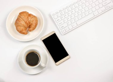 Üst ya da düz ekran cep telefonu, kahve fincanı, kruvasan, beyaz arkaplanda fotokopi alanı olan bilgisayar klavyesi. Sabah çalışma zamanı konsepti.