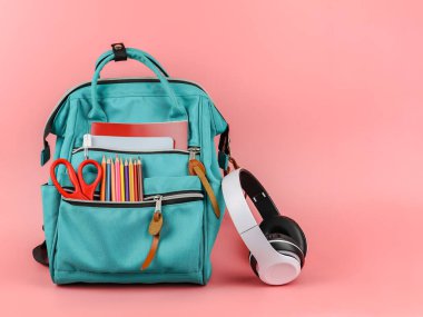 Okul gereçleriyle dolu sırt çantasının ön görüntüsü ve pembe arka planda fotokopi alanı olan beyaz kulaklıklar. Okula ve eğitim konseptine dönüş.