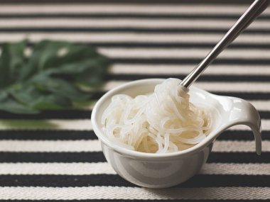 Metal çubuklar, siyah beyaz çizgili masa örtüsü arka planında kaldırılan beyaz kasedeki Konjac veya Shirataki şehriyelerinin ön görüntüsü.