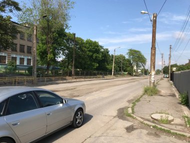 Gümüş rengi yeni araba, eski bir kasabanın çamurlu caddesinde asfalt yol boyunca gidiyor.