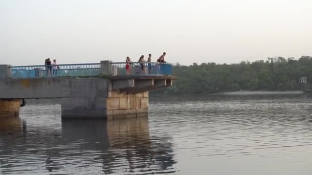 Вечірній літній краєвид біля річки в місті з видом на пірс, де діти отримують задоволення і стрибають у воду, яка заборонена і небезпечна. — стокове відео