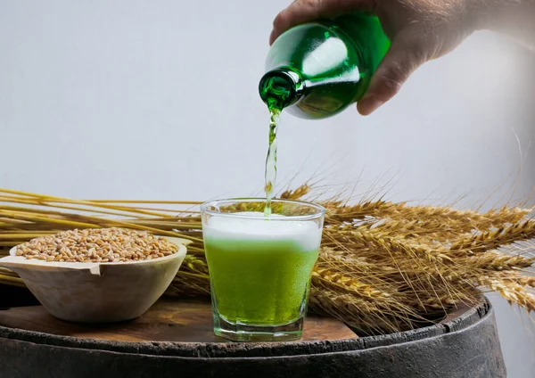 A man pours an original green beer from a green bottle into a small glass that is on a wooden oak barrel. The beer atmosphere is made up of wheat and its cob.