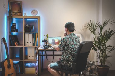 Yaratıcı, hippi ve modern çalışma alanında çalışan sakallı yakışıklı genç bir erkek. Laptop, kamera ve renkli ışıklar. Nüfuz Edicinin Ev Seti.