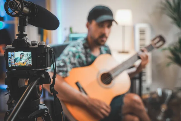 Behind the scene shot of a young man influencer making a video playing guitar. Very handsome man with beard shooting a video for social media talking directly to the camera. Influencer\'s work space.