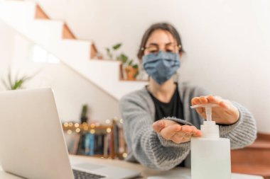 Coronavirus. Genç iş kadını evden izole edilmiş, koruyucu maske takıyor. Koronavirüs için karantinada olan kadın çalışırken dezenfektan jeli kullanıyor. Karantina. 