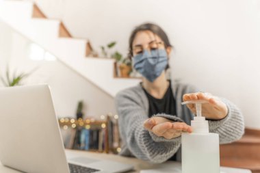 Coronavirus. Genç iş kadını evden izole edilmiş, koruyucu maske takıyor. Koronavirüs için karantinada olan kadın çalışırken dezenfektan jeli kullanıyor. Karantina. 