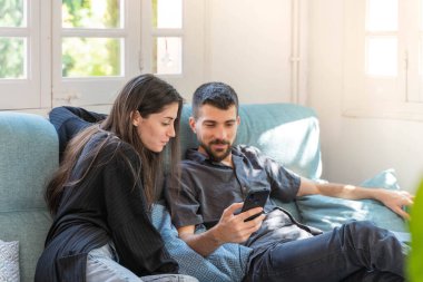 Coronavirus. Bir çift, Coronavirus sırasında evdeki karantinada kanepede oturuyor. Evdeki çift kanepede oturuyor, cep telefonuna bakıyor ve konuşuyor. - Eve. Evde kal.. 