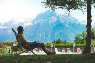 Genç iş adamı dizüstü bilgisayarla güzel ve harika manzaralı dağ geçmişi üzerinde çalışıyor. Yüzme havuzunun yanında çalışan bir iş adamı. Serbest çalışan biri. Gezgin. 