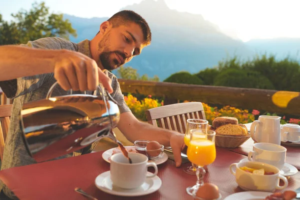 Man enjoying amazing breakfast table in nature during a sunny day with beautiful view. Healthy breakfast with toast, fruit slices, fresh juice, yoghurt, nuts and maple sirup. Homemade.