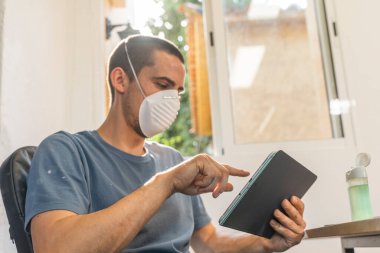 Coronavirus. Koronavirüs yüzünden karantinada olan bir adam koruyucu maske takıyor. Pencerenin yanındaki cep telefonuyla izole edilmiş halde evde. Görüntülü arama. Cep telefonuyla video konferansı. Karantina.