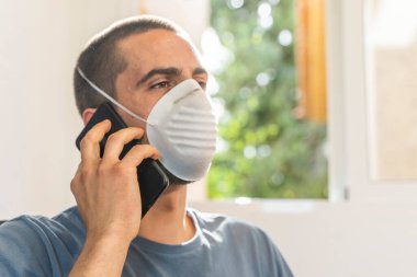 Coronavirus. Koronavirüs yüzünden karantinada olan bir adam koruyucu maske takıyor. Pencerenin yanındaki cep telefonuyla izole edilmiş halde evde. Görüntülü arama. Cep telefonuyla video konferansı. Karantina.