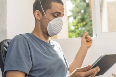 Coronavirus. Koronavirüs yüzünden karantinada olan bir adam koruyucu maske takıyor. Pencerenin yanındaki cep telefonuyla izole edilmiş halde evde. Görüntülü arama. Cep telefonuyla video konferansı. Karantina.