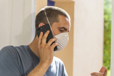 Coronavirus. Koronavirüs yüzünden karantinada olan bir adam koruyucu maske takıyor. Pencerenin yanındaki cep telefonuyla izole edilmiş halde evde. Görüntülü arama. Cep telefonuyla video konferansı. Karantina.