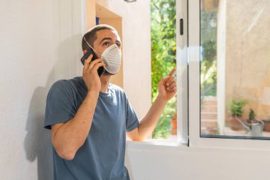 Coronavirus. Koronavirüs yüzünden karantinada olan bir adam koruyucu maske takıyor. Pencerenin yanındaki cep telefonuyla izole edilmiş halde evde. Görüntülü arama. Cep telefonuyla video konferansı. Karantina.