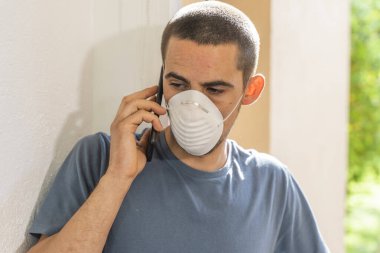 Coronavirus. Koronavirüs yüzünden karantinada olan bir adam koruyucu maske takıyor. Pencerenin yanındaki cep telefonuyla izole edilmiş halde evde. Görüntülü arama. Cep telefonuyla video konferansı. Karantina.
