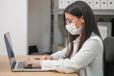 Bilgisayar ve yüz maskesi olan küçük bir şirkette çalışan bir iş kadını. Küçük bir şirket çalışıyor ve iş yapıyor. Küçük şirket ve takım çalışması konsepti. Küçük ofis. Karantina sırasında evde çalışıyorum. Coronavirüs.