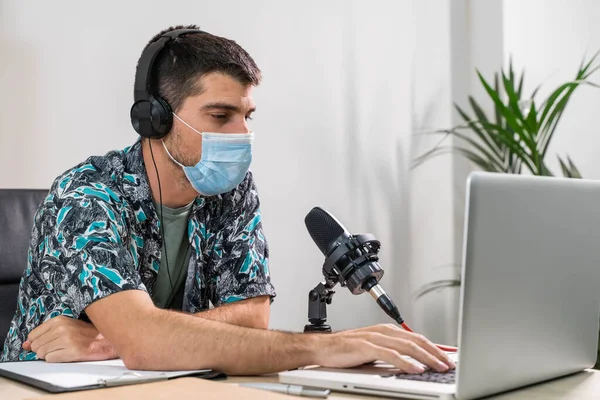 Man working as radio host at radio station sitting in front of microphone. Young radio host moderating a live show for radio with face mask. Working at home. Coronavirus.