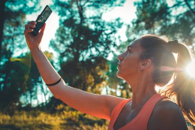 Ormanda akıllı telefonunu kullanarak yürüyen güzel bir genç kız. Genç, mutlu, çekici koşucu kadın, kır yollarında koşuyor... doğa geçmişi sağlıklı yaşam tarzı konseptinde neşeli koşuyor..