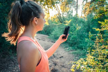 Ormanda akıllı telefonunu kullanarak yürüyen güzel bir genç kız. Genç, mutlu, çekici koşucu kadın, kır yollarında koşuyor... doğa geçmişi sağlıklı yaşam tarzı konseptinde neşeli koşuyor..