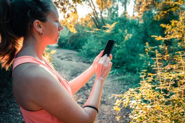 Ormanda akıllı telefonunu kullanarak yürüyen güzel bir genç kız. Genç, mutlu, çekici koşucu kadın, kır yollarında koşuyor... doğa geçmişi sağlıklı yaşam tarzı konseptinde neşeli koşuyor..