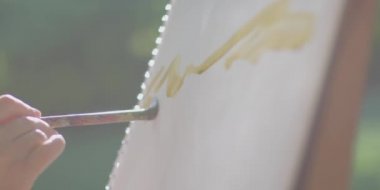 Bir sanatçının açık havada yaptığı resmin yakın görüntüsü. Ressamın ellerinde fırça, suluboya boya, yaratıcılık. Sanatçı çiçek bahçesindeki bir sehpada manzara resmediyor ve güneşli bir günde.