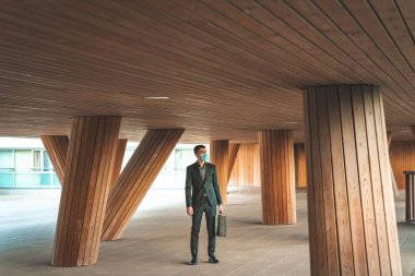 Kendinden emin ve başarılı. Surat maskesi takan yakışıklı genç işadamı arka planda ofis binasıyla dışarıda dikiliyor.