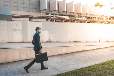 Kendinden emin ve başarılı. Arkaplanda ofis binasıyla dışarıda dolaşırken cerrahi maske takan yakışıklı genç bir işadamı ve bir evrak çantası..