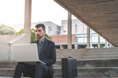 Dışarıda merdivenlerde oturan yakışıklı genç iş adamı dizüstü bilgisayarıyla çalışıyor. Mesafe çalışıyor. Takım elbiseli yalnız bir adam. Metin için alanı kopyala.