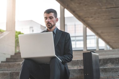Dışarıda merdivenlerde oturan yakışıklı genç iş adamı dizüstü bilgisayarıyla çalışıyor. Mesafe çalışıyor. Takım elbiseli yalnız bir adam. Metin için alanı kopyala.