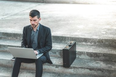 Dışarıda merdivenlerde oturan yakışıklı genç iş adamı dizüstü bilgisayarıyla çalışıyor. Mesafe çalışıyor. Takım elbiseli yalnız bir adam. Metin için alanı kopyala.