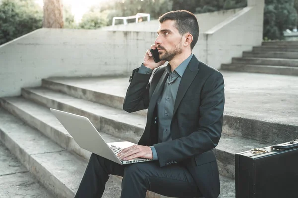 Handsome young businessman sitting on the stairs outdoors working with his laptop while talking on his phone. Distance working. Isolated man on a suit.