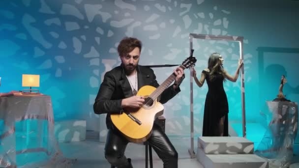 Joven músico tipo en una chaqueta negra toca y canta en una guitarra, detrás de él la chica pone en estudio de arte. — Vídeo de stock