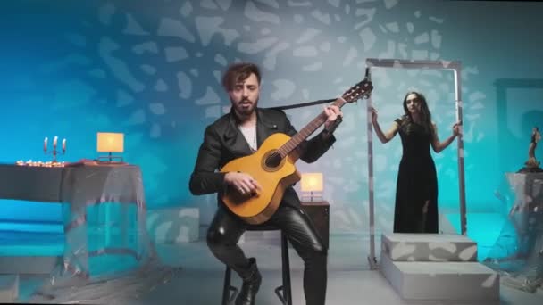 Giovane musicista ragazzo in una giacca nera gioca e canta su una chitarra, dietro di lui la ragazza posa in studio d'arte — Video Stock