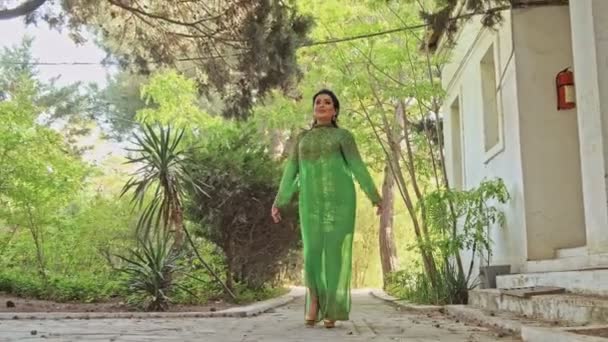 Loop in de tuin. Een brunette vrouw in een groene jurk loopt langs de bomen — Stockvideo