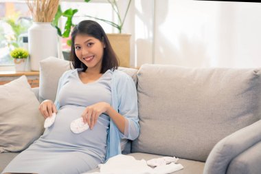 Genç ve çekici hamile Asyalı kadın bebeği için evde kıyafet hazırlıyor. Mutlu anne internetten akıllı telefon ıslatma kullanıyor..