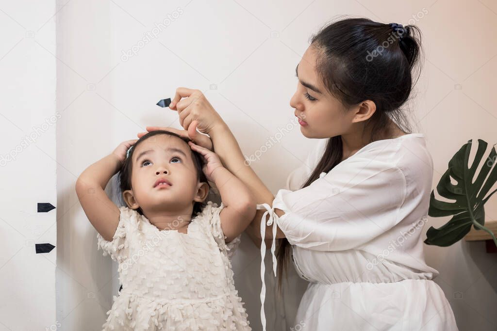 Young attractive Asian Mom measuring her daughter's tall and mark it on the wall.