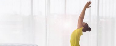 Sarı tişörtlü genç Asyalı bayan sabah uyandıktan sonra yatak odasında yoga pozu veriyor..
