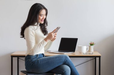 Genç mutlu Asyalı kadın ofiste akıllı telefon kullanıyor..