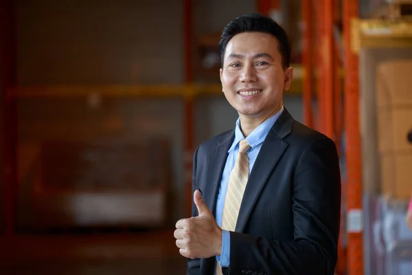 Portrait of asian business man owner company in black formal suit standing with confident showing thumps up, doing good job, on warehouse factory background, Business Investor concept.
