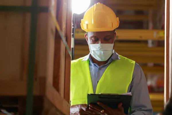 Young black African worker wearing protective face mask working in factory warehouse. Black man checking stock, New normal after covid 19 pandemic crisis. Logistic industry concept.