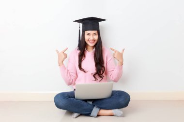 Genç ve çekici Asyalı bir kadın dizüstü bilgisayarı ile oturmuş mezun olmuş bir şapka takıyor, online eğitim kursu, yurtdışında burs konsepti..