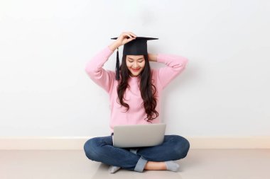 Genç ve çekici Asyalı bir kadın dizüstü bilgisayarı ile oturmuş mezun olmuş bir şapka takıyor, online eğitim kursu, yurtdışında burs konsepti..
