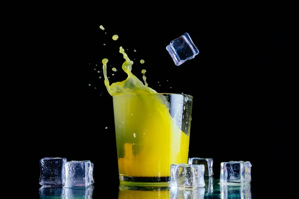 Splashes of orange juice in a clear glass with ice cubes on a dark background