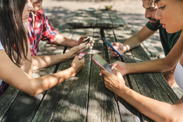 Young people using smartphone over an old wooden table in the courtyard. Friends using cell phones on Sunday in the summer. Technology and social media concept.