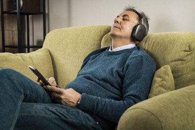 Kanepede oturan, kablosuz kulaklıklı, akıllı telefonlu, gözleri kapalı evde müzik dinleyen son sınıf öğrencisi. Rahatlama, akış, teknoloji, yaşam tarzı, rahatlık, rahatlık ve refah kavramı.