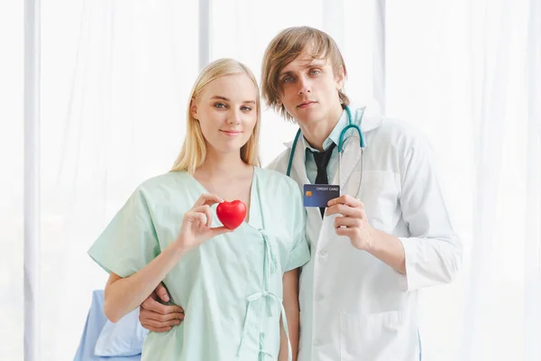 A female patient is holding a red heart-shaped ball and a doctor is happily carrying a credit card in hand. Concept of cardiology and healthcare people in a hospital. Good health and credit card.
