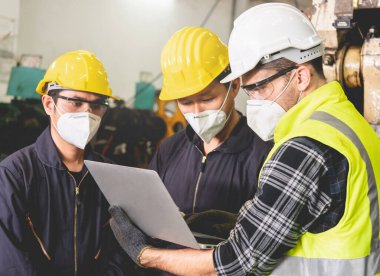 Başlıklı fabrika mühendisi, fabrikanın içindeki iş detaylarını tartışmak için dizüstü bilgisayar tutuyor. Sert şapkalı endüstriyel mühendis ekibi fabrikada çalışıyor..